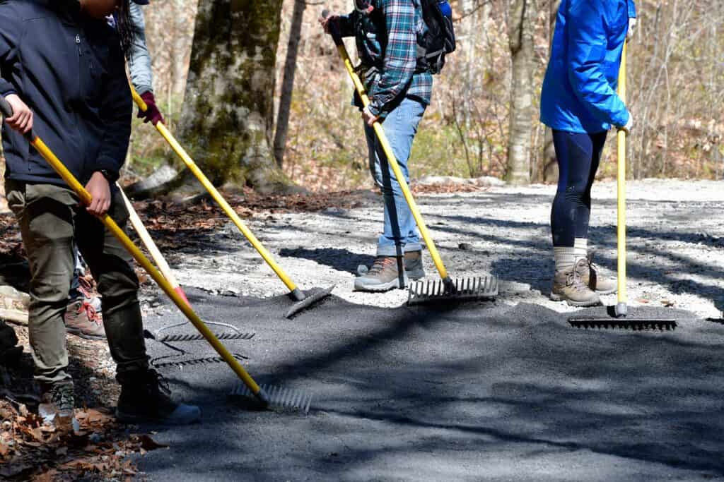 Workers leveling fresh concrete on a forest trail with rakes and tools.